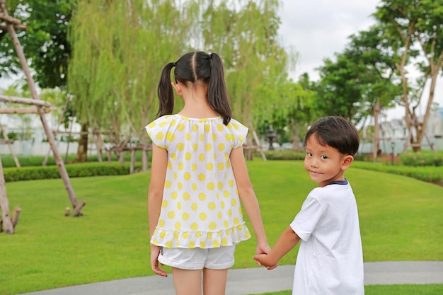 귀여운 아시아 소년과 소녀가 정원에서 손을 잡고 있는 동안 어린 소년이 카메라를 보고 있는 동안 녹색 공원에 있는 아시아 어린이