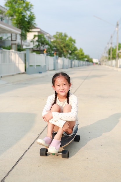 かわいいアジアの小さな女の子が通りでスケート ボードに座ってリラックスします。