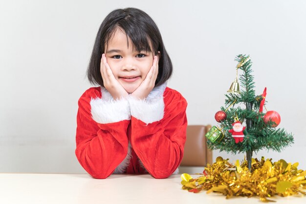 A cute Asian little girl in a red Santa Claus dress decorating Christmas tree on Christmas eve