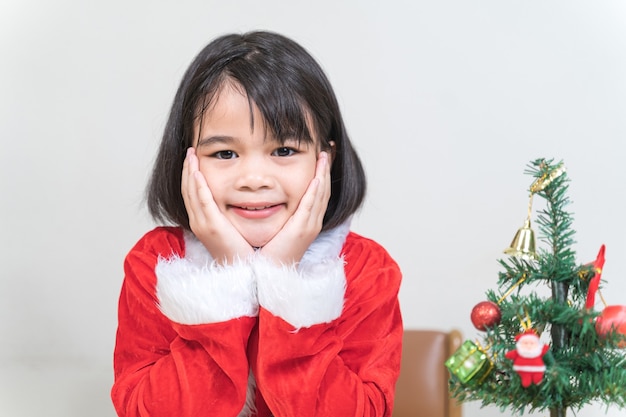 A cute Asian little girl in a red Santa Claus dress decorating Christmas tree on Christmas eve