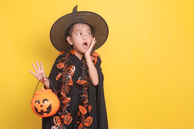 Cute asian little girl wearing witch costume holding pumpkin bucket for candy and showing surprise e