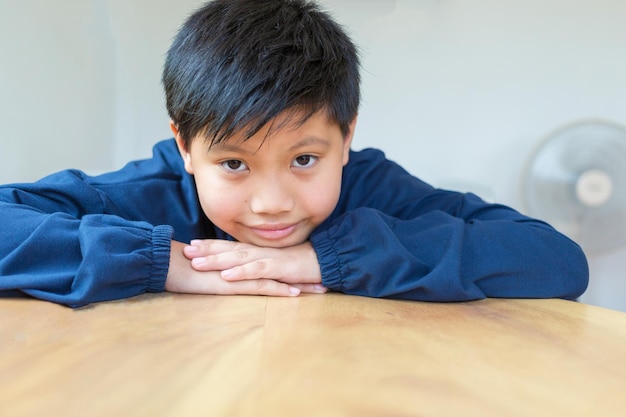 Cute Asian little kid boy with smiling looking in camera happily resting chins on handson table