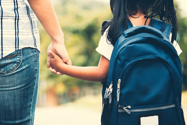 Cute asian pupil girl with backpack holding her mother hand and going to school