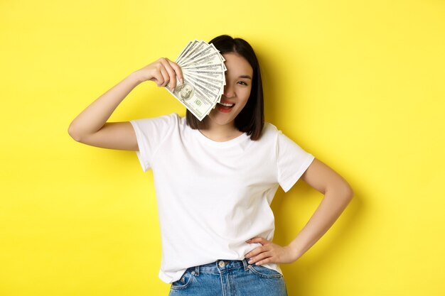 お金の後ろに顔を隠し、満足しているカメラをのぞき、現金を稼ぎ、黄色の上に立っているかわいいアジアの女性。