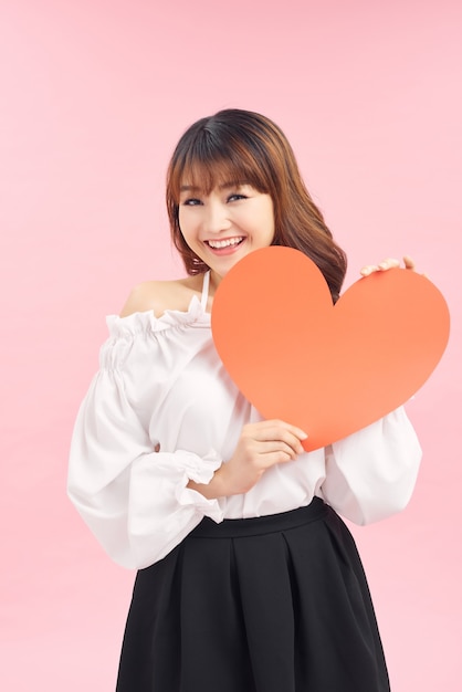 Cute asian woman holding red heart isolated on pink