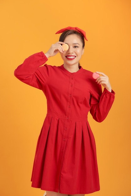 Cute Asian woman in red dress holding macaron over orange background