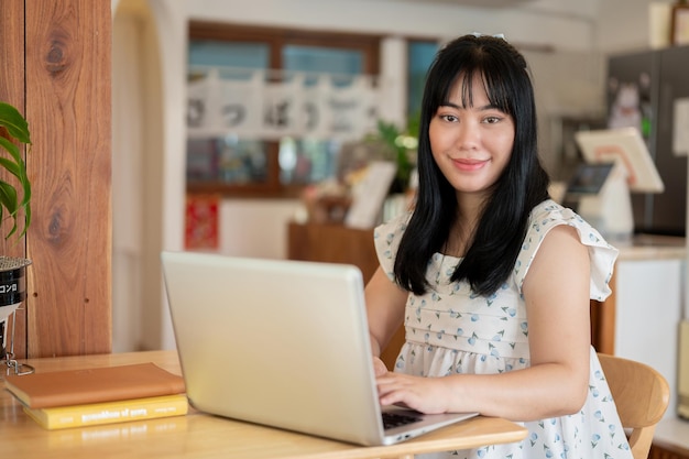 Милая азиатская женщина сидит за столом в кафе со своим ноутбуком, улыбаясь в камеру.