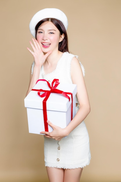 Cute Asian woman smile and holding white gift box Pretty girl model wearing white beret and natural makeup on beige background Cosmetology beauty and spa wellness Plastic surgery