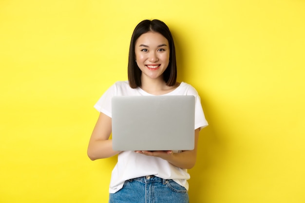 Cute asian woman studying on laptop and smiling, standing in white t-shirt and jeans against yellow background.