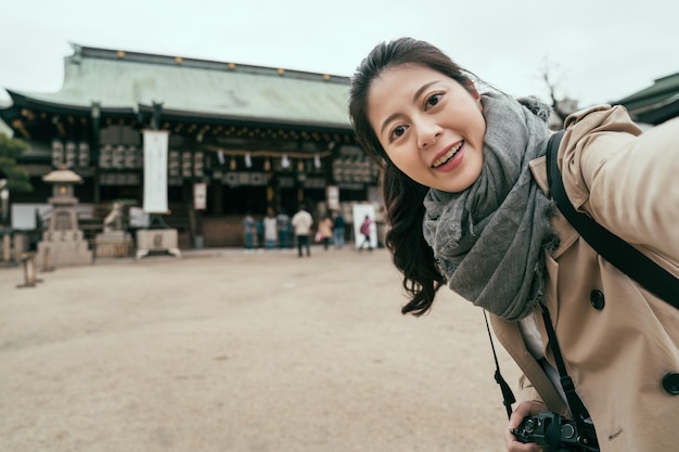 cute asian woman traveler taking selfie at temple in tenmangu osaka. female tourist recording while sightseeing in japan. girl travel photographer make self photo in shrine face camera smiling