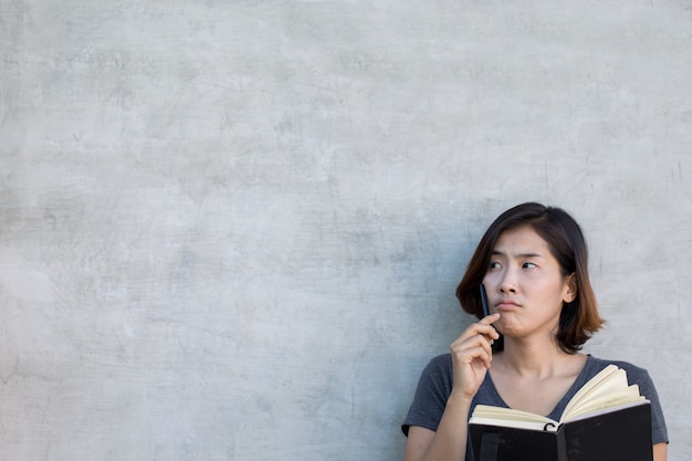 Foto le donne asiatiche carine stanno pensando qualcosa con il suo libro su sfondo grigio