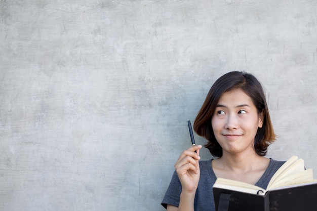 Foto le donne asiatiche carine stanno pensando qualcosa con il suo libro su sfondo grigio