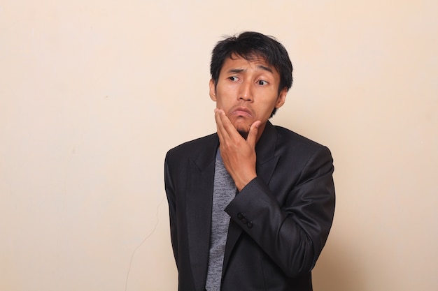 Cute Asian young man with thinker expression is pulling down his lips, wearing a suit with