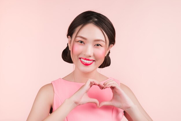 Cute asian young woman with fashion makeup gesture hand making heart shape isolated on pink.