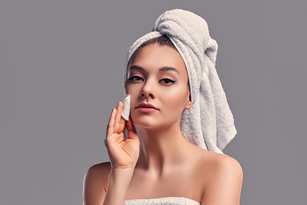 Cute attractive girl with a towel on her head rubs her face with a cotton pad with lotion isolated on a gray background. Skin care concept.