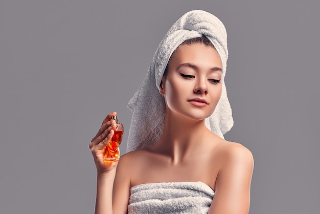 Cute attractive girl with a towel on her head uses perfume isolated on a gray background. Skin care concept. Spa treatments, cosmetology, make-up.
