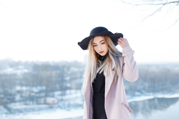 Cute attractive young woman in a chic black hat in a vintage elegant pink coat in a black knitted dress posing on the backdrop of a winter river on a sunny winter day. Glamor blonde girl.