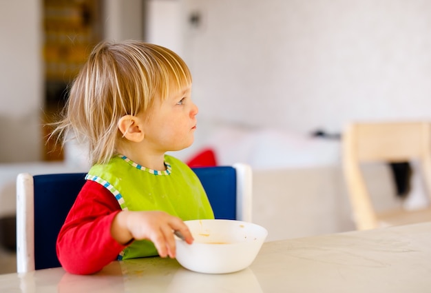 Foto bambino sveglio 1,4 anni seduto sulla sedia per bambini alti e mangiare verdura da solo nella cucina bianca.