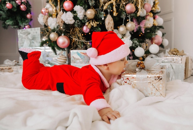 A cute baby 6 months old in a Santa costume lies on a blanket near the Christmas tree. christmas 2022