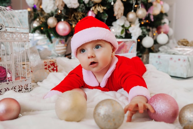 A cute baby 6 months old in a Santa costume lies on a blanket near the Christmas tree. christmas 2022