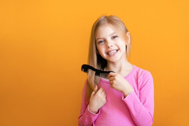 Cute baby, baby girl 6-8 years old combs her soft blond hair medium length comb and smiles on an orange wall