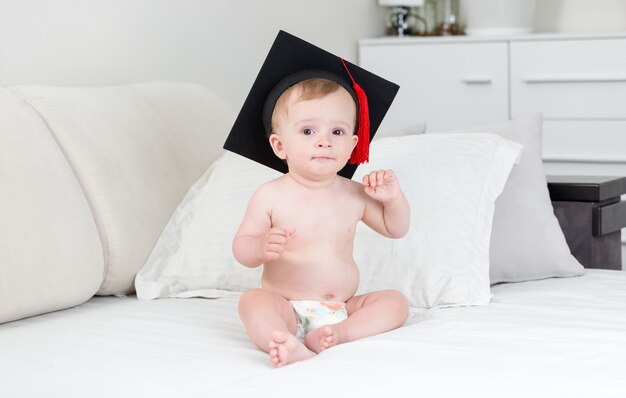 Cute baby boy in academic black hat sitting on bed