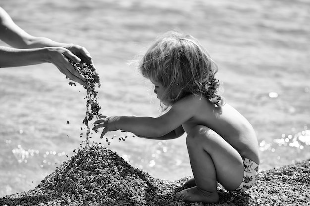 Cute baby boy child with curly blond hair sit on beach and plays with pebble on blue sea background ...