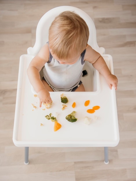 Cute baby boy in highchair eating vegetables alone