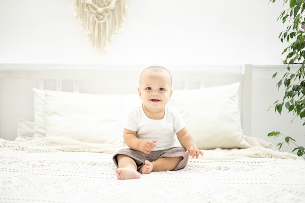 Cute baby boy is sitting on a bed with white linens in the bedroom of the house a child's portrait the child is looking at the camera