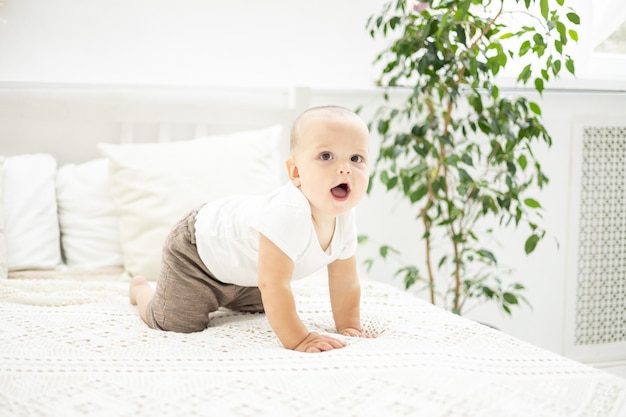 Photo cute baby boy is sitting on a bed with white linens in the bedroom of the house a child's portrait the child is looking at the camera