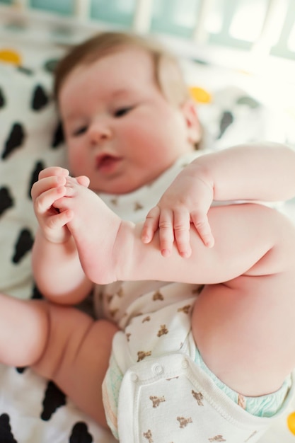 ベッドに横になって足で遊ぶかわいい男の子赤ちゃんの部屋で晴れた朝