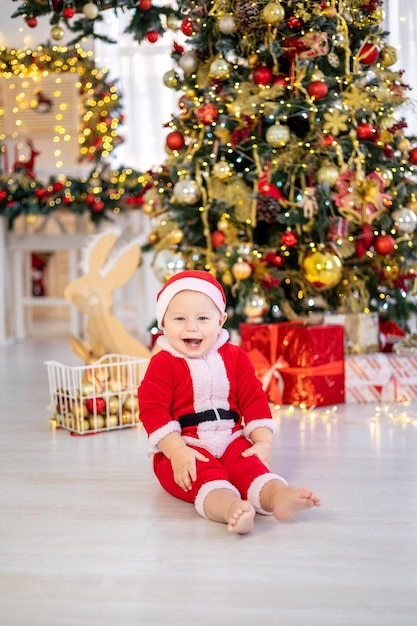 A cute baby boy in a Santa costume is sitting with Christmas tree toys under a festive Christmas