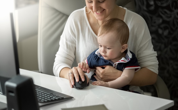 Cute baby boy sitting on mothers lap and playing with computer mouse