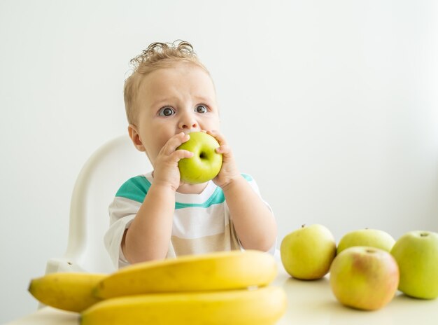 Cute baby boy sitting at the table in child chair eating apple on white kitchen