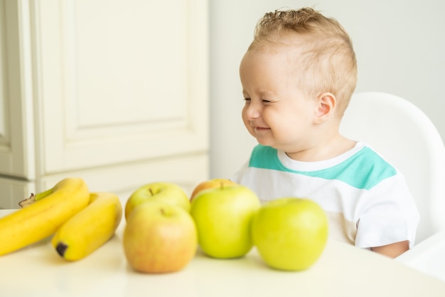 Cute baby boy sitting at the table in child chair eating apple on white kitchen