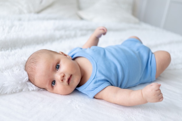 Cute baby boy three months old in a blue bodysuit on a white bed at home.