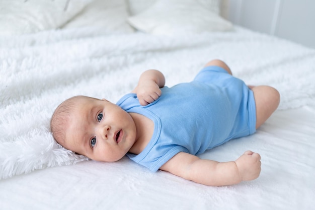 Cute baby boy three months old in a blue bodysuit on a white bed at home