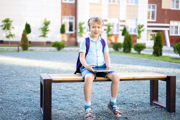 校庭の遠隔学習のベンチに座っているワイヤレスヘッドフォンとタブレットのかわいい男の子