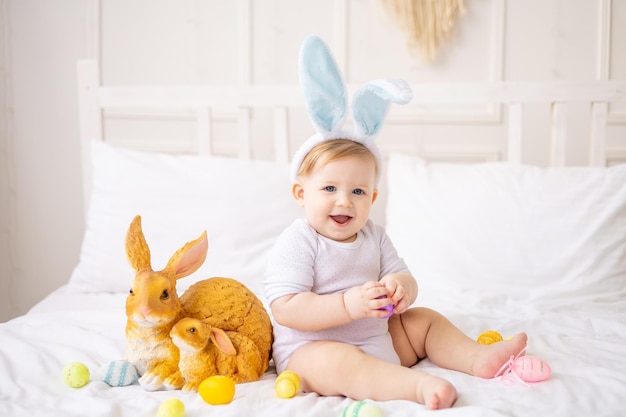 Cute baby boy with bunny ears and colorful eggs on a white bed at home playing little blonde baby with a rabbit happy easter concept