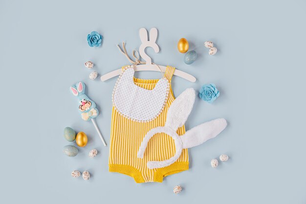 Cute baby costume Easter bunny. Knitted bodysuit with fluffy  bunny ears, Easter eggs and candy on blue background. Holiday concept. Flat lay, top view