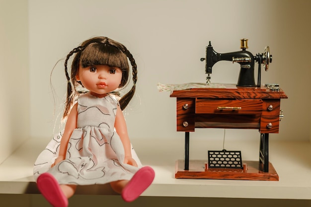 Cute baby doll with white clothing sitting next to the sewing machine toy and looking at camera Cute doll photoshoot