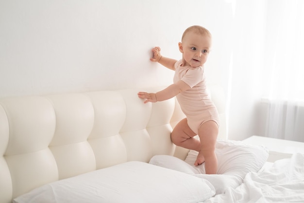 Cute baby girl in bodysuit stand holding bed frame learning to walk on white bedding on bed