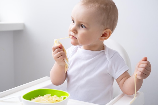 cute baby girl eating spaghetti pasta sitting in baby chair on white kitchen