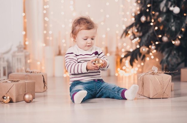 Cute baby girl holding Christmas ball sitting on floor over Christmas tree