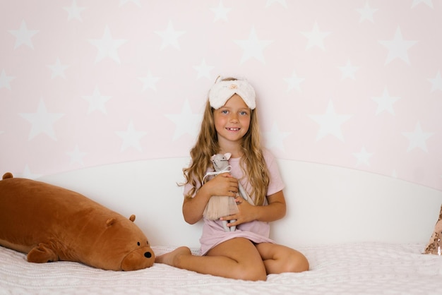 Милая девочка обнимает мягкую игрушку, сидящую на кровати в спальне
