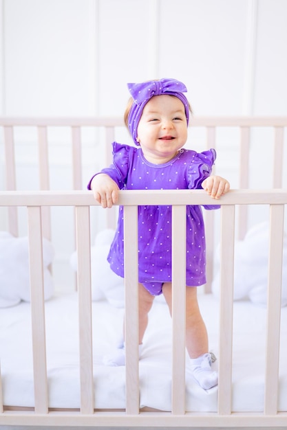 Cute baby girl is standing in a crib and holding on to the side in lilac clothes and laughing the concept of children's goods