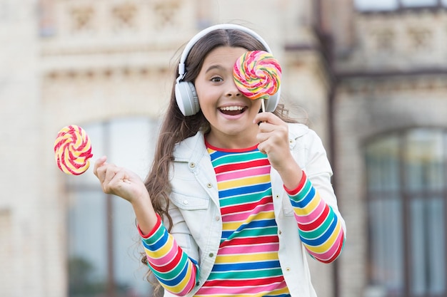 Cute baby girl kid have fun at schoolyard healthy food child has music break girl in headphones small girl in wireless headset with lollipop back to school modern education with new technology