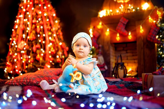 Cute baby girl in pajama sitting on checkered plaid near Christmas tree and fireplace and garlands