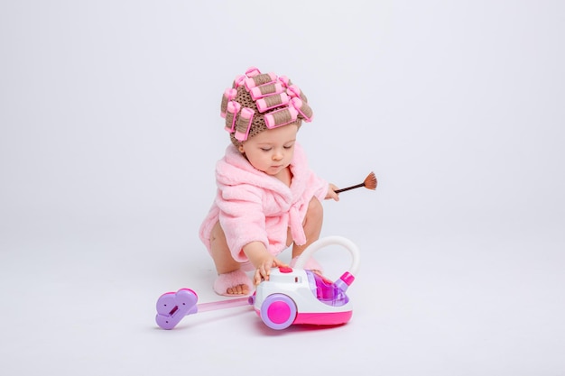 장난감 진공 청소기가 달린 분홍색 테리 천을 입은 귀여운 여자 아이는 흰색 배경에 격리되어 있습니다