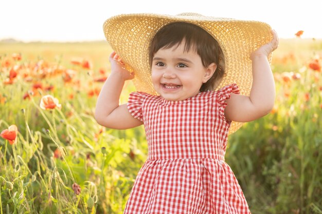 Cute baby girl in red dress and straw hat on field of poppies at summer sunset.